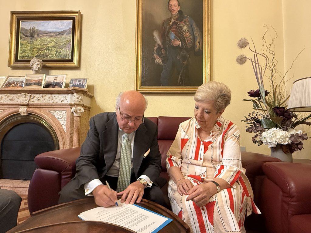 Dos personas sentadas firmando un documento en una sala con cuadros y chimenea