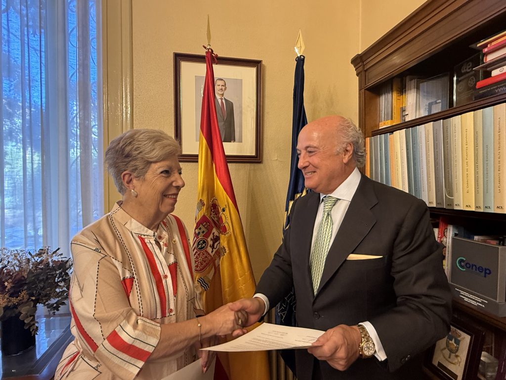Dos personas se estrechan la mano con un documento, junto a una bandera de España, en un despacho