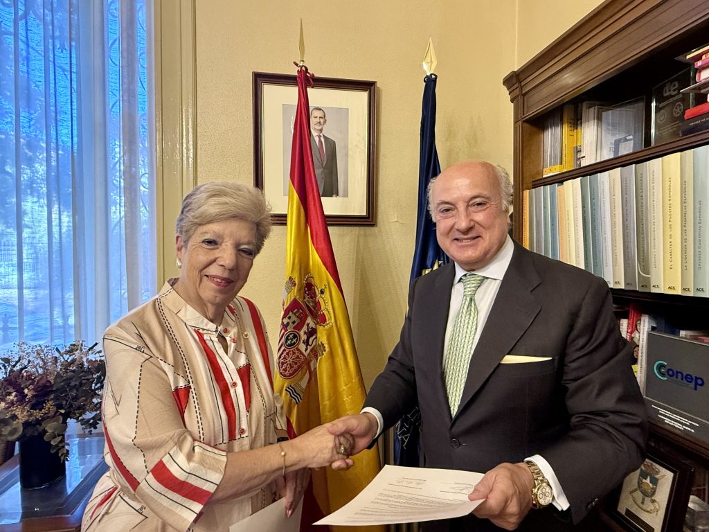 Dos personas se estrechan la mano con un documento, junto a una bandera de España, en un despacho